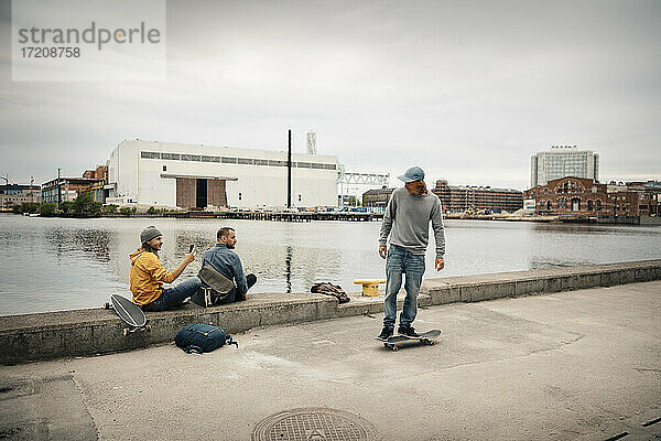 Mann auf Skateboard stehend  während männlicher Freund durch Smartphone fotografierend am Kanal sitzt