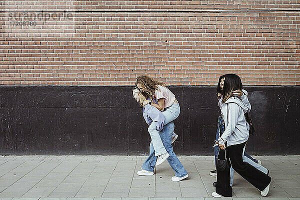 Glückliche Freundinnen tragen Huckepack  während sie an der Wand auf dem Fußweg gehen