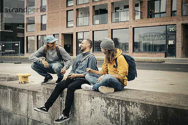 Glückliche männliche Freunde lachen beim Sitzen auf Stützmauer gegen Gebäude