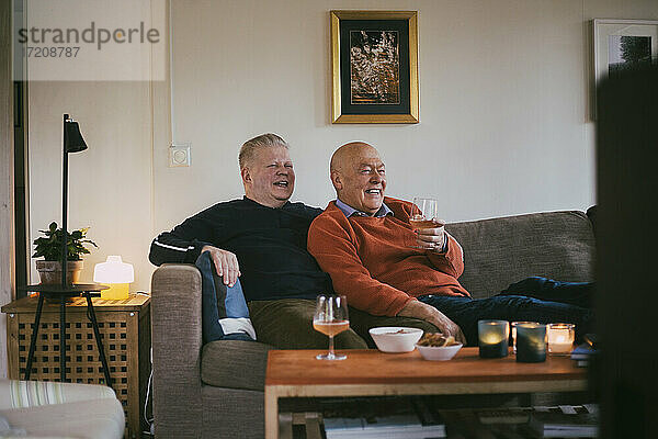 Lächelndes homosexuelles Paar mit Weinglas beim Fernsehen zu Hause