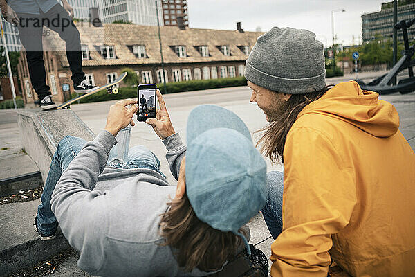 Freunde fotografieren durch Handy während männlicher Skater mit Skateboard auf Stützmauer