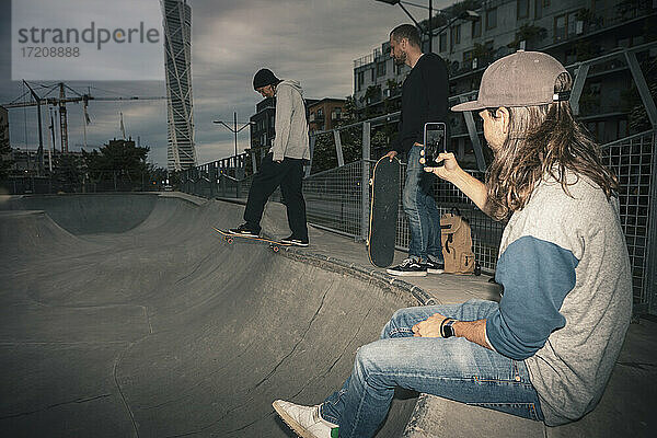 Männlicher Skateboarder skatet  während ein Freund mit seinem Smartphone im Park fotografiert