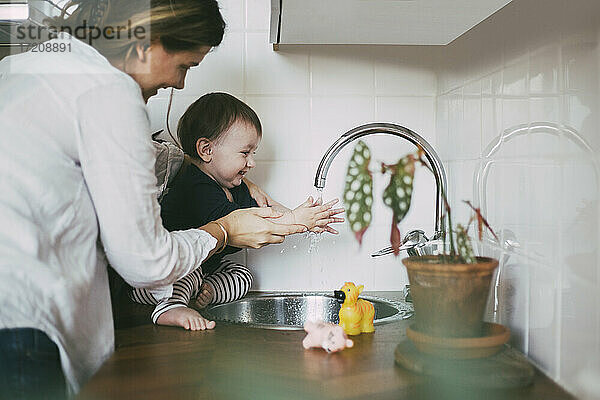 Lächelnde Mutter hilft Baby Mädchen Hände waschen unter Waschbecken in der Küche