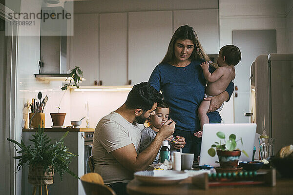 Vater füttert Sohn  während Mutter mit Baby-Mädchen in der Küche steht