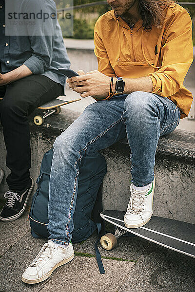 Niedriger Ausschnitt eines Mannes  der ein Smartphone benutzt und neben einem männlichen Freund auf einer Stützmauer sitzt