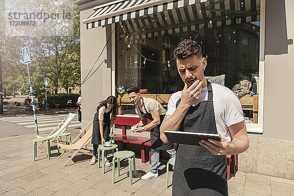 Männlicher Unternehmer  der ein digitales Tablet benutzt  während Kollegen im Hintergrund vor einem Café arbeiten