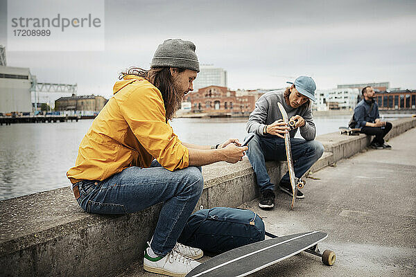 Männliche Freunde mit Smartphone und Skateboard sitzen auf Stützmauer