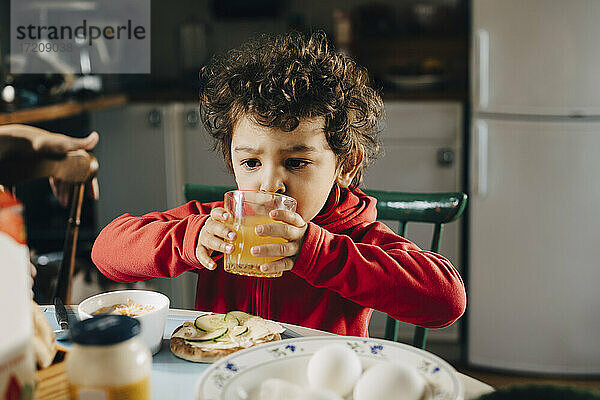 Netter Junge trinkt Saft beim Sitzen am Tisch während des Frühstücks