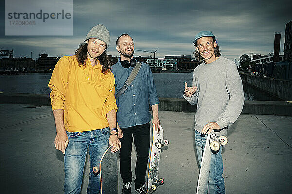 Porträt von lächelnden männlichen Freunden mit Skateboards gegen Kanal in der Abenddämmerung