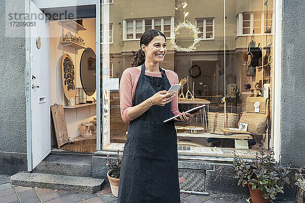 Lächelnde Unternehmerin mit Smartphone und digitalem Tablet vor dem Geschäft