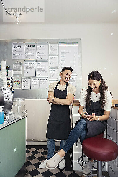 Weibliche Unternehmerin mit Smartphone  während ein männlicher Kollege mit verschränkten Armen in einem Café herumsteht