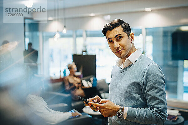 Porträt von Geschäftsmann mit Smartphone  während Kollegen im Hintergrund im Büro arbeiten