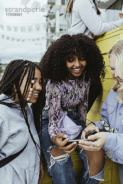 Lächelnde Freundinnen  die ein Smartphone benutzen  während sie auf einer Kiste im Freien sitzen