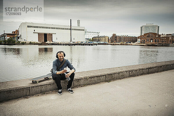 Lächelnder Mann mit Smartphone sitzt auf Stützmauer gegen Kanal