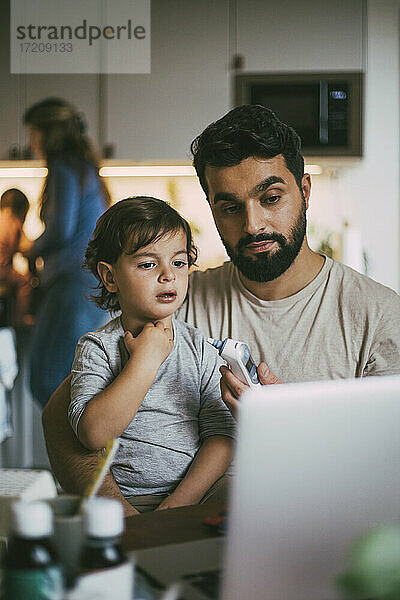 Vater hält Otoskop  während er mit Sohn während Online-Konsultation zu Hause sitzt