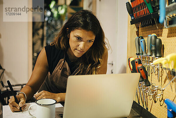 Weibliche Unternehmerin mit Laptop beim Schreiben auf Papier in einer Reparaturwerkstatt