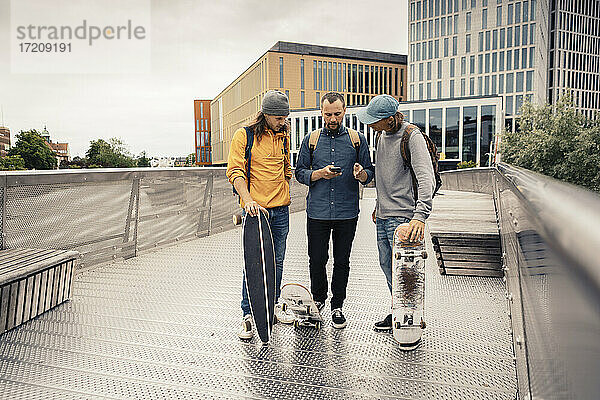 Freunde teilen Smartphone mit männlichen Skatern  während sie auf einer Brücke stehen