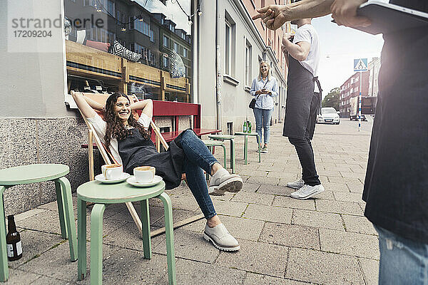 Lächelnde weibliche Besitzerin mit Händen hinter dem Kopf sitzt auf Liegestuhl außerhalb Kaffee-Shop