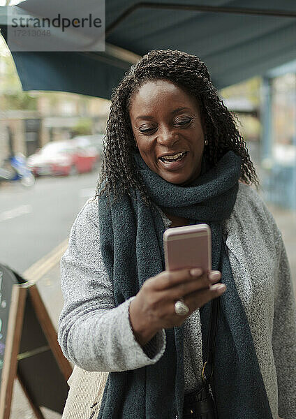 Glückliche Frau in Schal mit Smartphone auf dem Bürgersteig