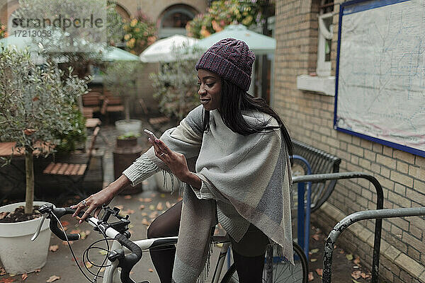 Junge Frau mit Smartphone auf dem Fahrrad vor einem Straßencafé