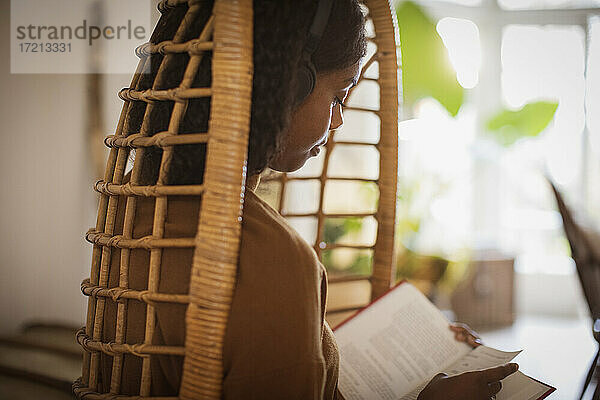 Serene junge Frau mit Kopfhörer lesen Buch in Rattan Stuhl