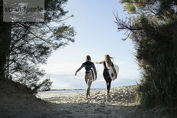 Junge Surferinnen laufen mit Surfbrettern auf einem sonnigen Strandweg