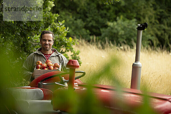 Portrait glücklicher Mann mit frisch geernteten Äpfeln am Traktor im Obstgarten