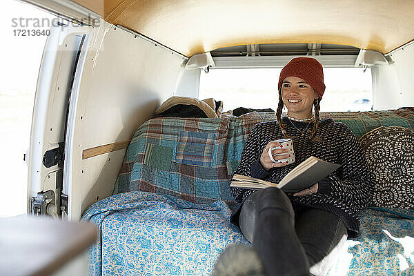 Glückliche junge Frau entspannt mit Buch und Tee im Wohnmobil