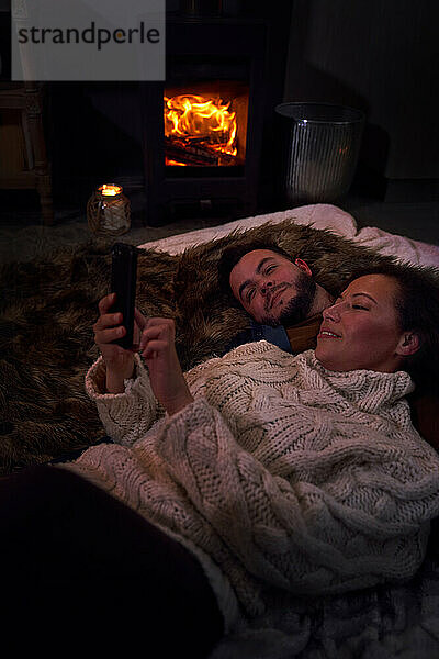 Romantisches Paar mit Smartphone am gemütlichen Kaminfeuer