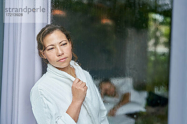 Neugierige Frau im Bademantel schaut aus regnerischem Fenster