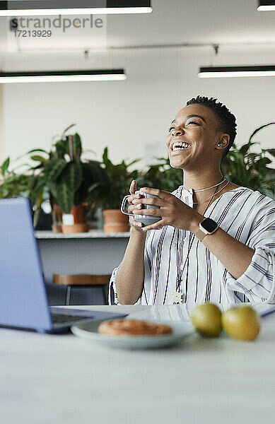 Glückliche Geschäftsfrau mit Kaffee lachend am Laptop im Büro