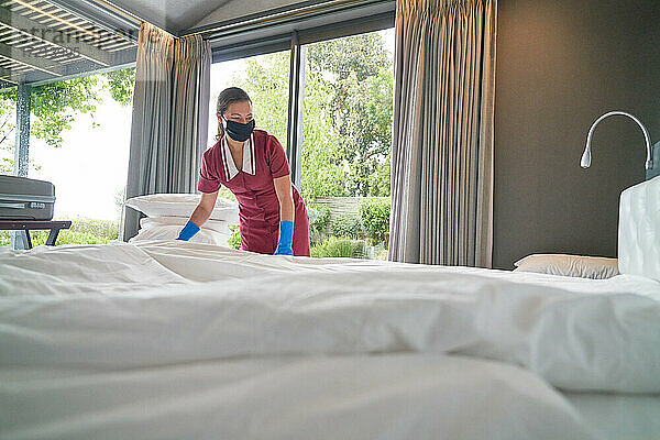 Weibliches Hotelzimmermädchen in Gesichtsmaske macht Bett im Hotelzimmer