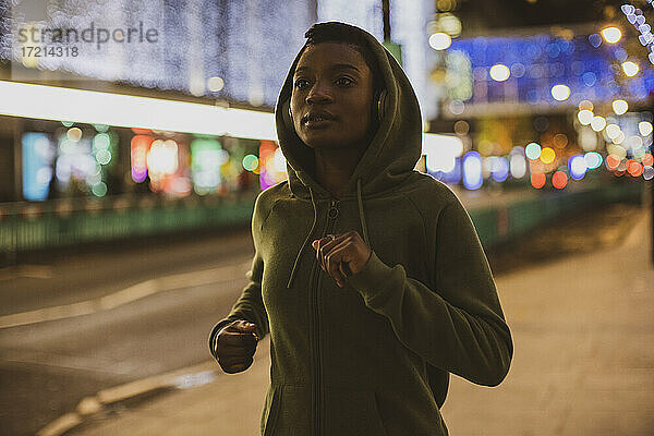 Junge Frau im Kapuzenpulli joggen auf Stadtstraße mit Lichtern in der Nacht