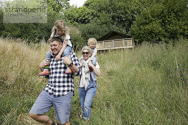Glückliche Eltern tragen Töchter auf der Schulter im hohen Gras unterhalb der Hütte