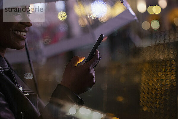 Junge Frau mit Smartphone mit Regenschirm in der Nacht