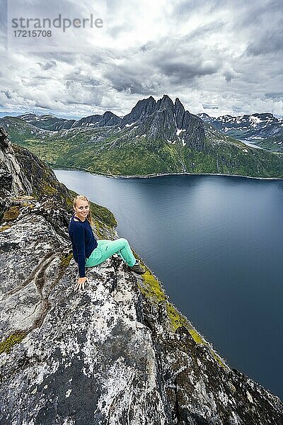 Junge Wanderin sitzt an Felsklippe  Abgrund zum Fjord  Ausblick vom Berg Barden  Senja  Norwegen  Europa