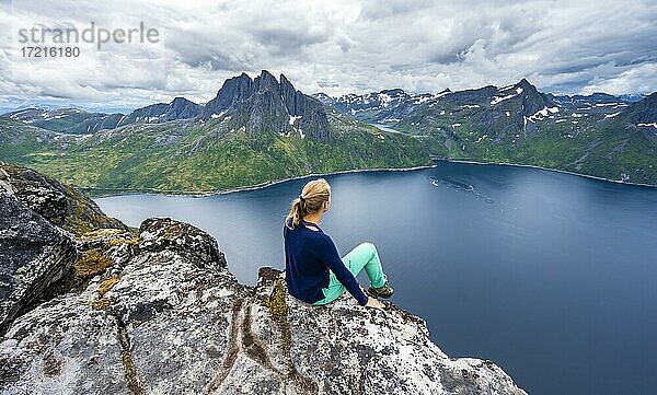 Junge Wanderin sitzt an Felsklippe  Abgrund zum Fjord  Ausblick vom Berg Barden  Senja  Norwegen  Europa