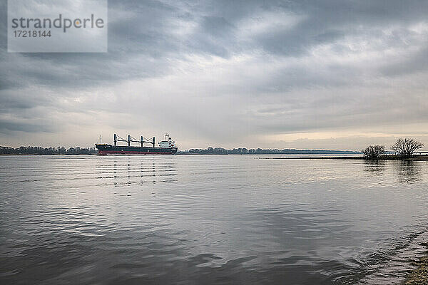 Frachtschiff ( BC Vanessa ) steuert auf der Elbe mit auflaufend Wasser den Hamburger Hafen an. Blick vom Elbstrand in Hamburg Rissen  es ist ein windstiller  bewölkter Winter-Nachmittag