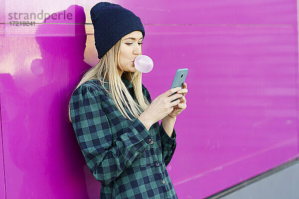 Frau im karierten Kleid macht Kaugummi-Blase  während mit Smartphone vor der Wand