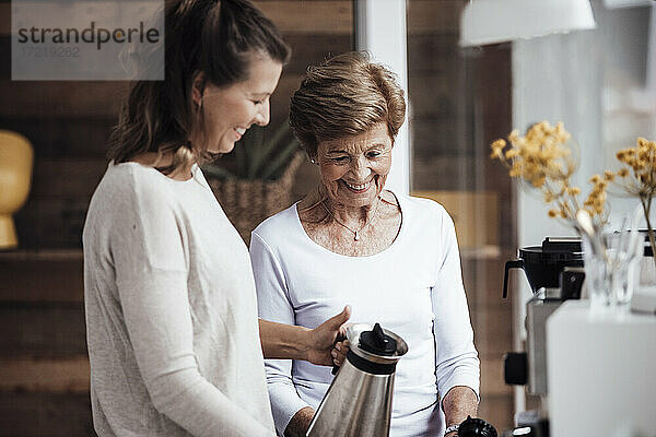 Lächelnde junge Frau  die zu Hause bei ihrer Großmutter einen Wasserkocher hält