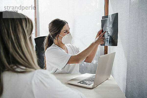 Eine Ärztin mit Gesichtsschutzmaske zeigt einem Patienten am Schreibtisch in einer Klinik ein Röntgenbild