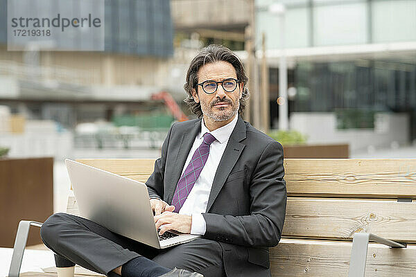 Nachdenklicher männlicher Fachmann  der einen Laptop benutzt  während er auf einer Bank im Büropark sitzt