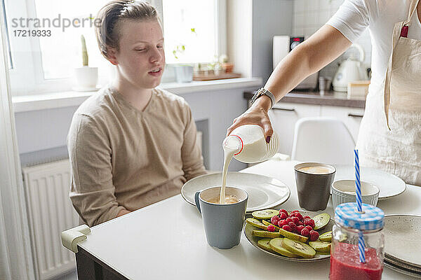 Frau gießt Milch in Tasse für Sohn im Teenageralter am Esstisch in der Küche