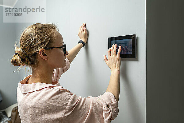 Junge Frau  die ein Hausautomatisierungsgerät benutzt  während sie zu Hause an der Wand steht
