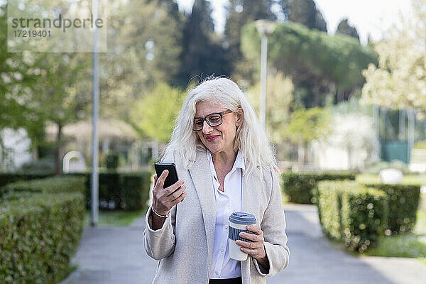 Lächelnde Frau  die ein Mobiltelefon benutzt und einen wiederverwendbaren Becher im Park hält