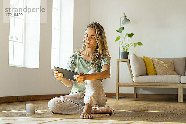 Frau in Loungewear schaut auf ein digitales Tablet  während sie im Wohnzimmer sitzt