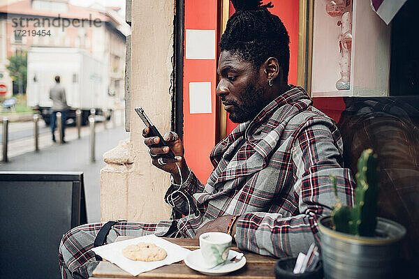 Modischer junger Mann mit Mobiltelefon in einem Straßencafé