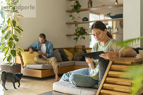 Junge Frau  die ein digitales Tablet benutzt  während ihr Freund zu Hause im Hintergrund ein Smartphone benutzt