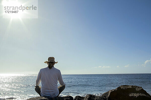 Älterer Mann sitzt auf einem Felsen vor dem Meer an einem sonnigen Tag