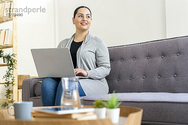 Frau  die wegschaut  während sie mit ihrem Laptop auf dem Sofa zu Hause sitzt